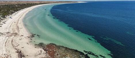 An aerial view of a beach with clear turquoise waters, a sandy shoreline, and sparse vegetation.