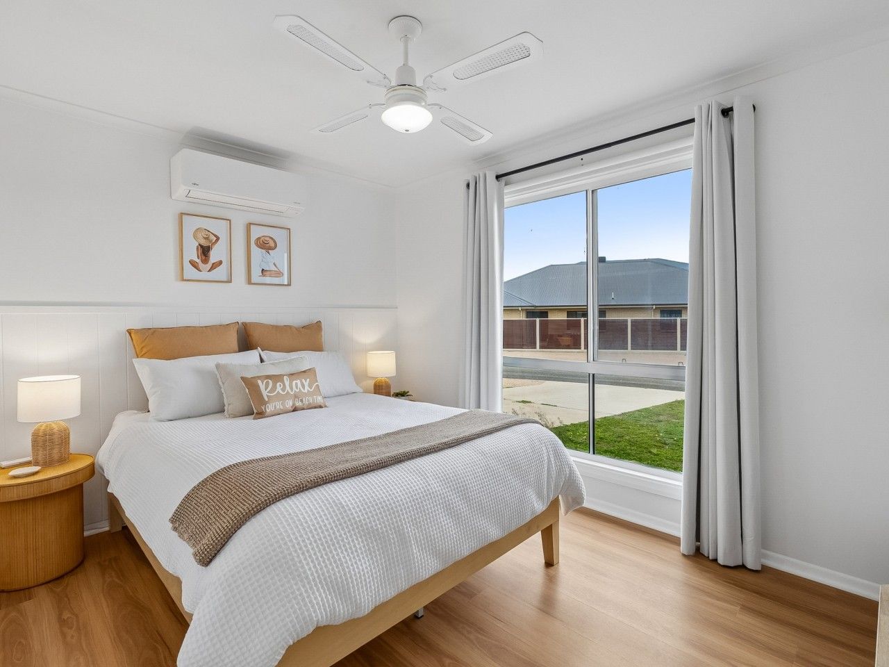 A bedroom with a bed, bedside lamps, a ceiling fan, a window with curtains, and a view of a building outside.