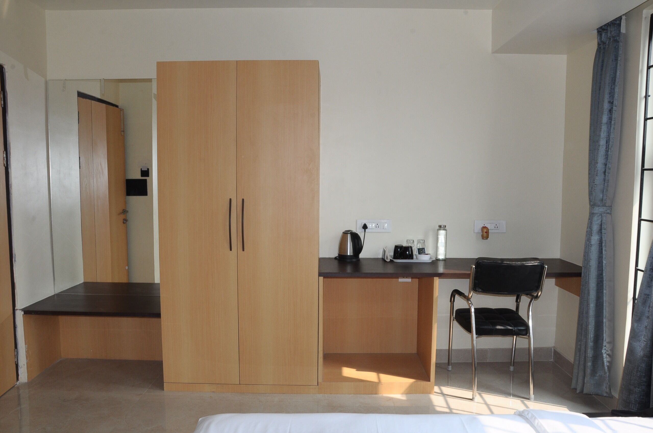 Une pièce de taille modeste, comprenant une armoire en bois, un petit bureau avec une chaise, une bouilloire et une prise électrique.