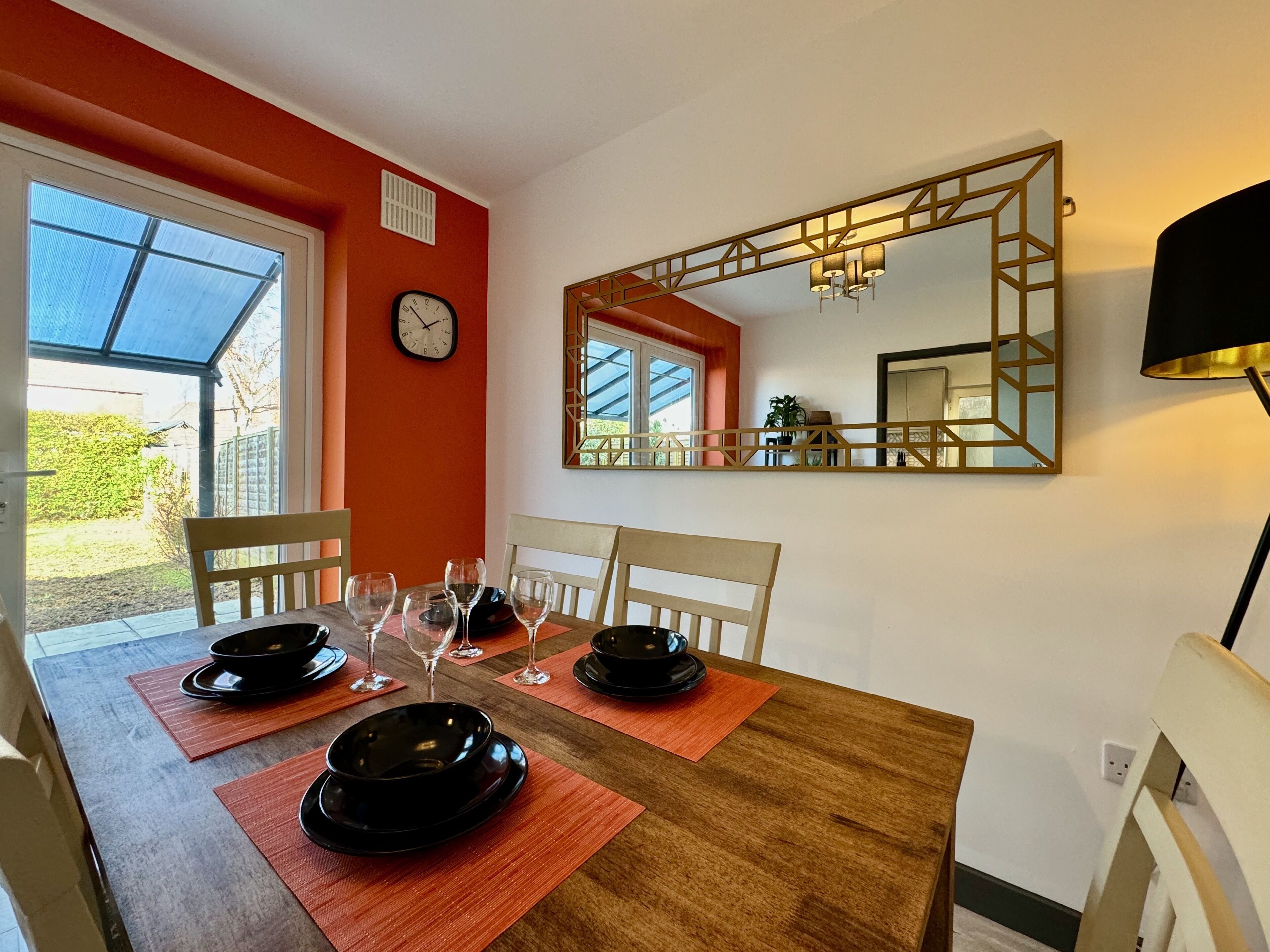 A dining area with a wooden table, black plates, and wine glasses. There is a clock on the wall and a mirror above the table.