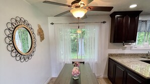 Un coin repas avec une table en bois, des chaises, un ventilateur de plafond et une vue par une fenêtre ornée de rideaux légers.