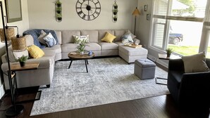 Un salon moderne avec un canapé d’angle, une table basse et un fauteuil. On y trouve une grande horloge murale et une fenêtre avec des stores.