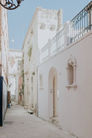 A narrow street with white buildings, arched doorways, and a metal railing on the upper floor.