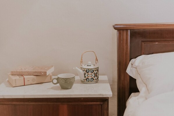 A neatly made bed with white linens, a bedside table with a white teapot and a green cup, and a stack of books on the table.