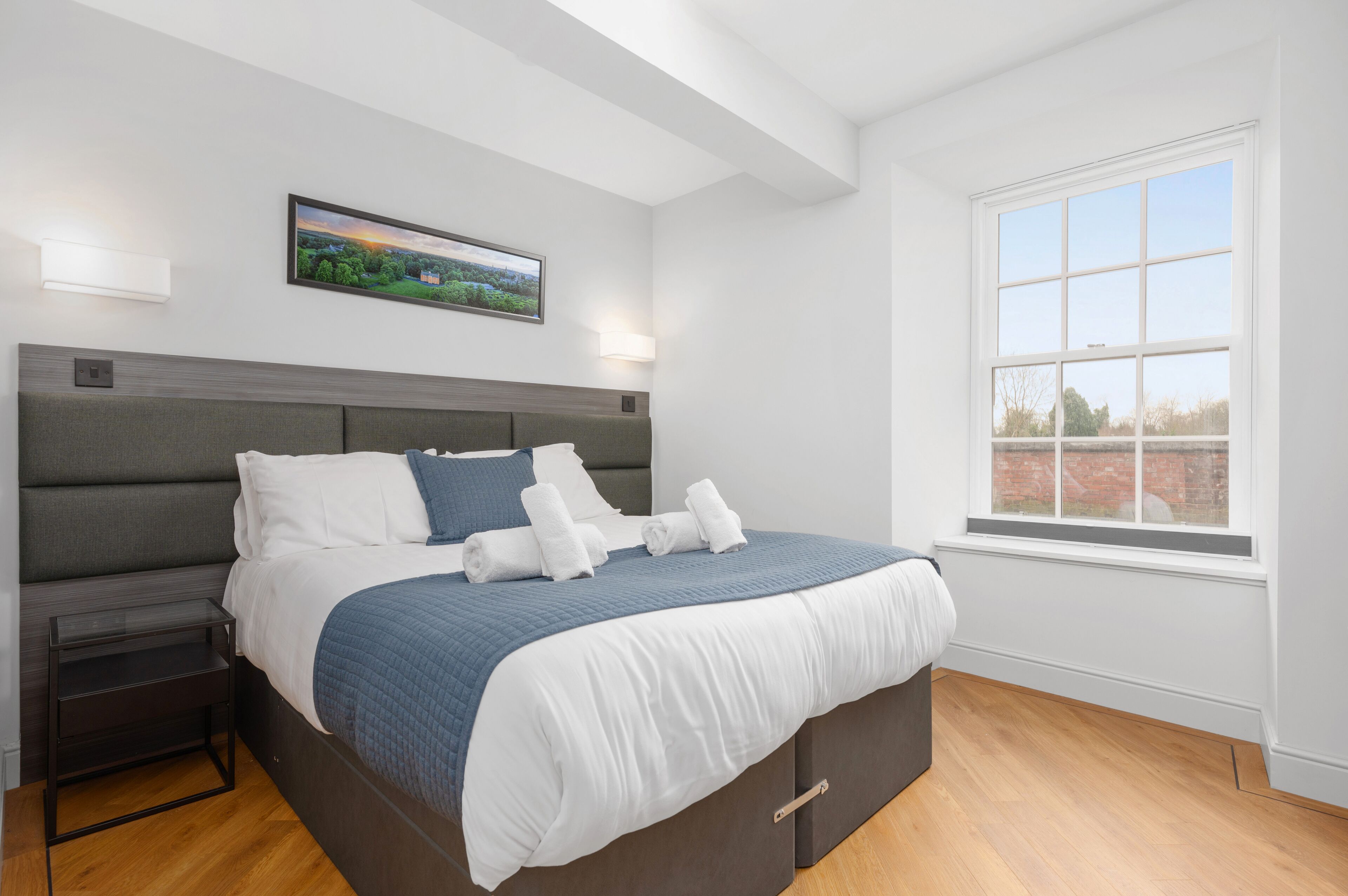 A modern bedroom with a large bed, a nightstand, a wall-mounted light, and a window with a view of the outside.