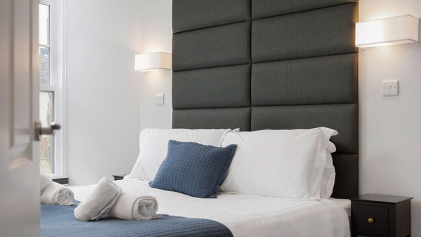 Un lit bien fait, avec une literie blanche et bleue, une tête de lit sombre et des lampes de chevet.