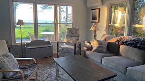 Un salón acogedor con sofá, sillón y mesa de centro. Amplias ventanas ofrecen vistas al exterior.