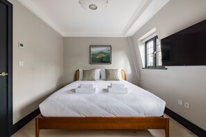 Ein ordentlich bezogenes Bett mit weißen Leinen und zwei Handtüchern, ein gerahmtes Landschaftsbild an der Wand und ein Fenster mit Jalousien auf der rechten Seite.