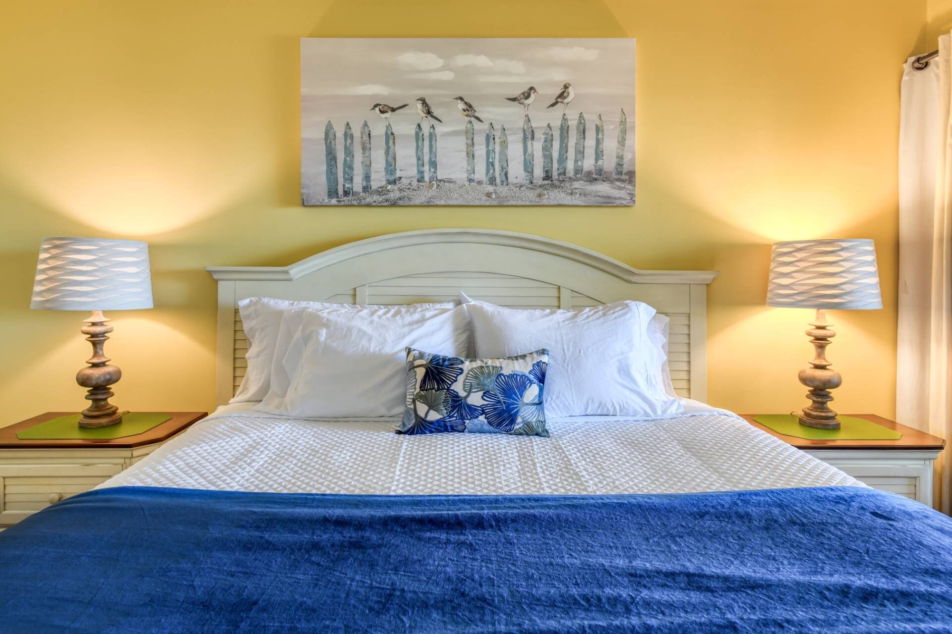 Un letto con biancheria bianca, una coperta blu e un cuscino con motivo blu e bianco. Ci sono due comodini con lampade e un quadro sopra il letto.