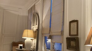 Una habitación con un ventanal grande, una silla de mimbre, una mesa con lámpara y un cuadro en la pared.