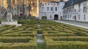 Un jardín formal con setos geométricos, un edificio histórico con una torre destacada y una estatua en primer plano.