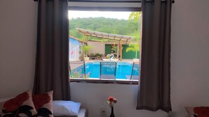 A room with a bed, a small table with a vase of flowers, and a window overlooking a swimming pool and outdoor area. - Pousada Sunflower (Pirenópolis)