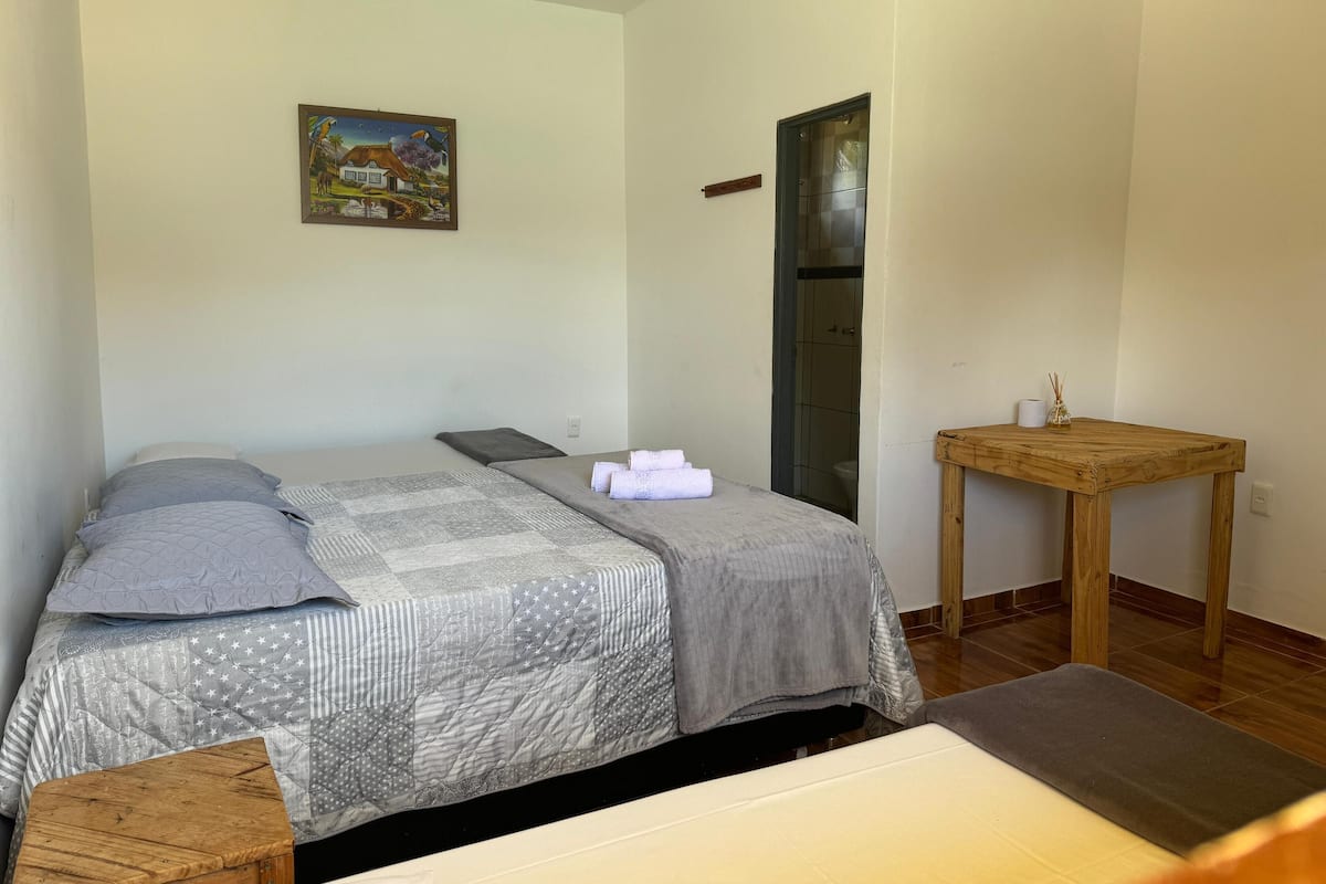 A bedroom with a bed, pillows, a quilt, a small wooden table, and a painting on the wall.