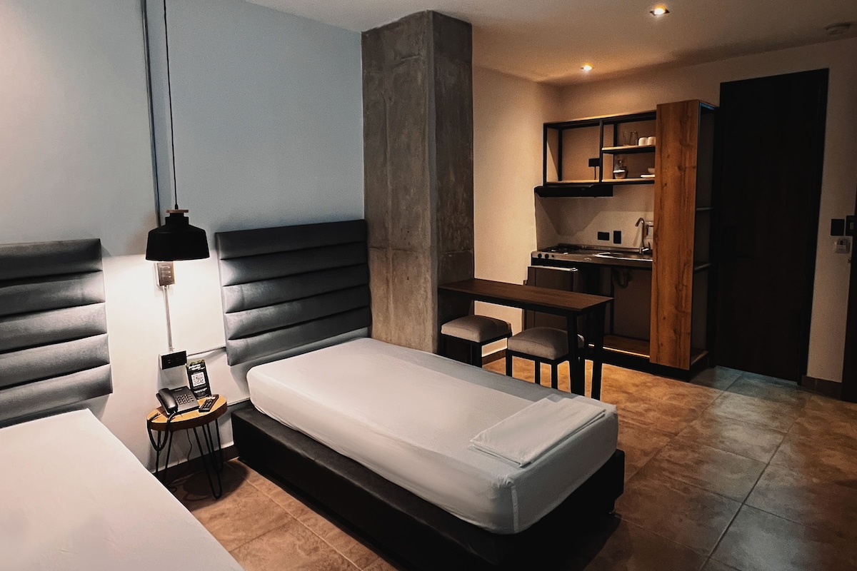 Een moderne hotelkamer met een bed, nachtkastje, lamp en een kitchenette.