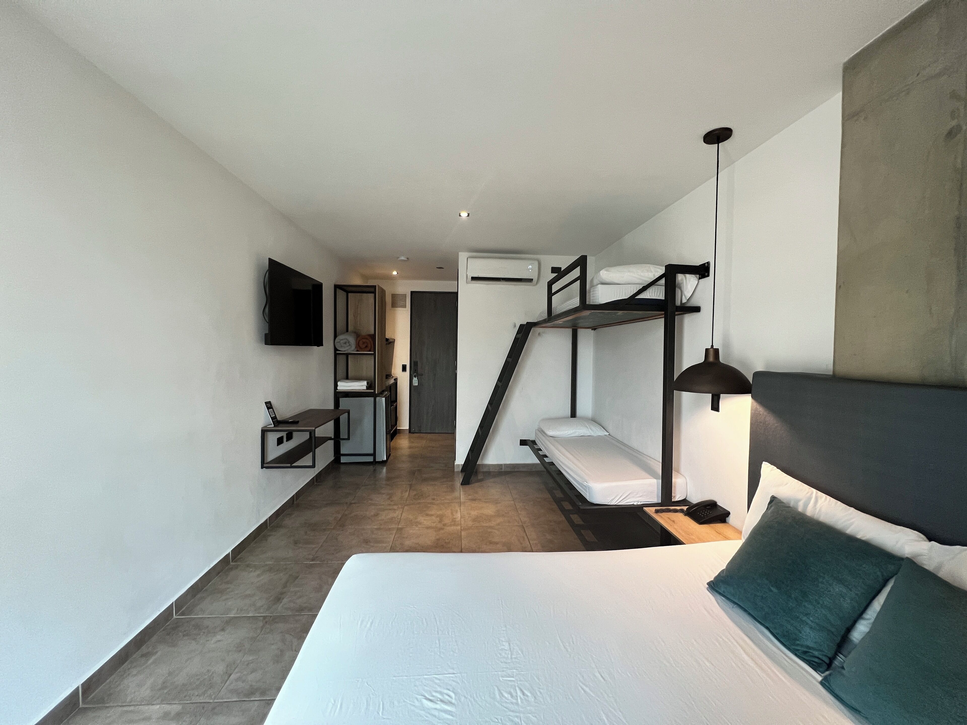 Habitación de hotel compacta con literas, un escritorio con silla, un televisor de pantalla plana en la pared y una lámpara de noche.