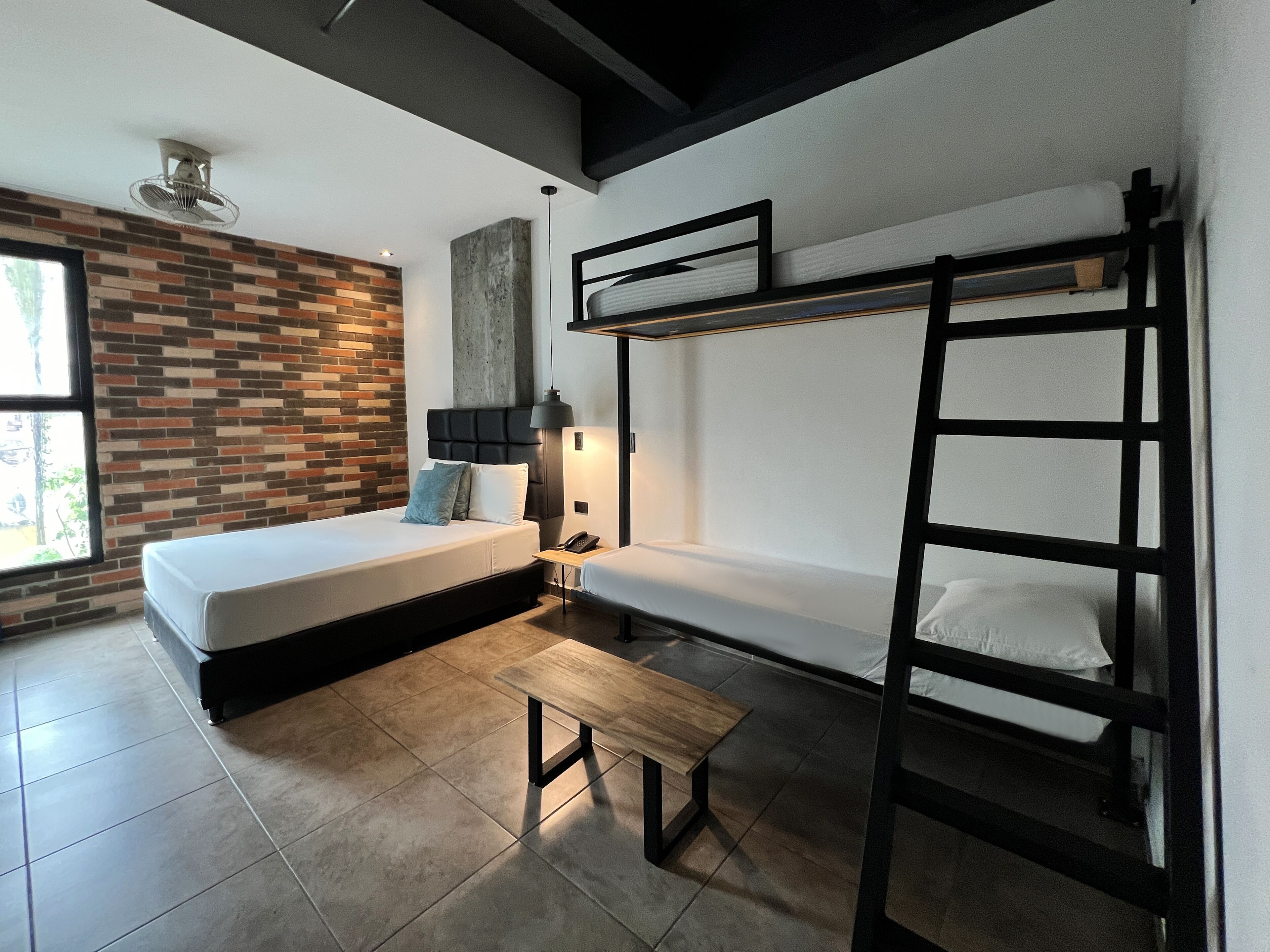 Ein modernes, kompaktes Zimmer mit einer Ziegelwand, einem Etagenbett, einem Nachttisch mit Lampe und einem Fenster für natürliches Licht.
