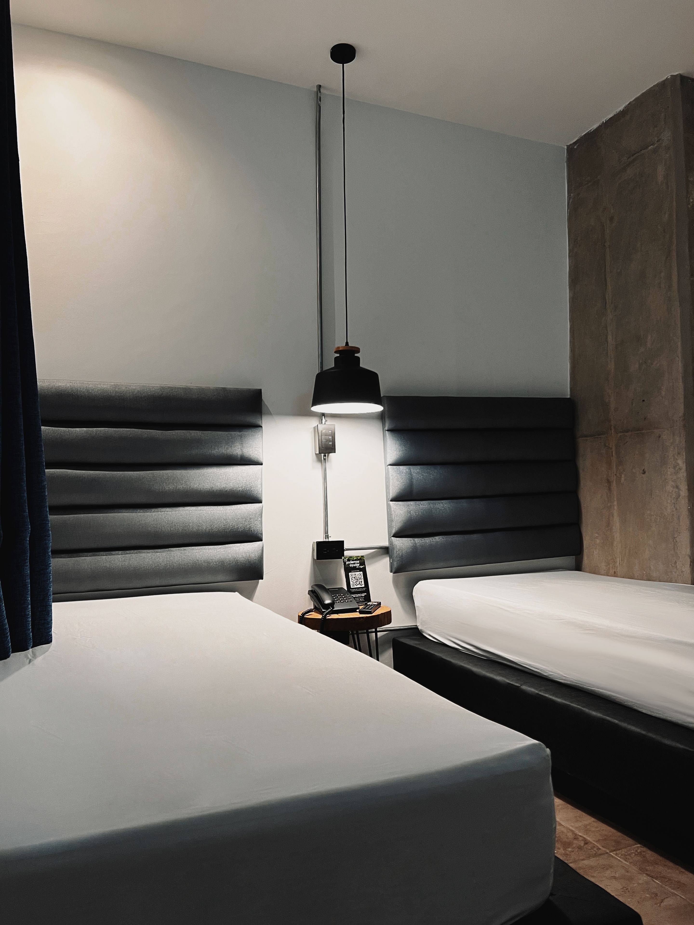 Habitación de hotel con dos camas, una mesita de noche con teléfono y lámpara, y una lámpara moderna.
