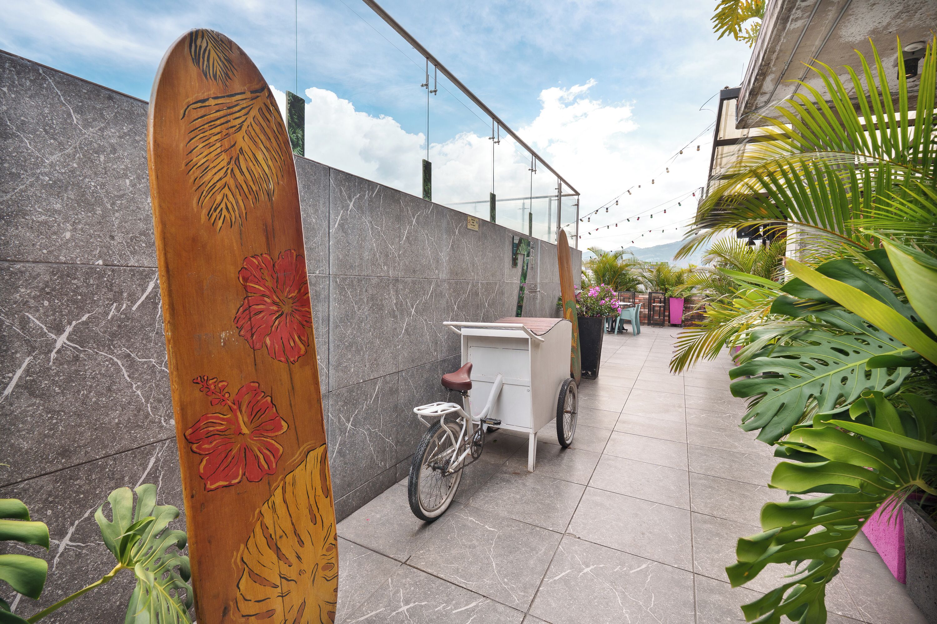 Ein Holzsurfbrett mit floralen Mustern, ein Fahrrad und ein Wagen stehen auf einer gefliesten Terrasse mit Pflanzen und einem Gebäude im Hintergrund.