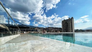 Une piscine sur le toit, avec une eau d’un bleu cristallin, entourée d’une balustrade en verre, et offrant une vue sur le paysage urbain et les montagnes en arrière-plan.