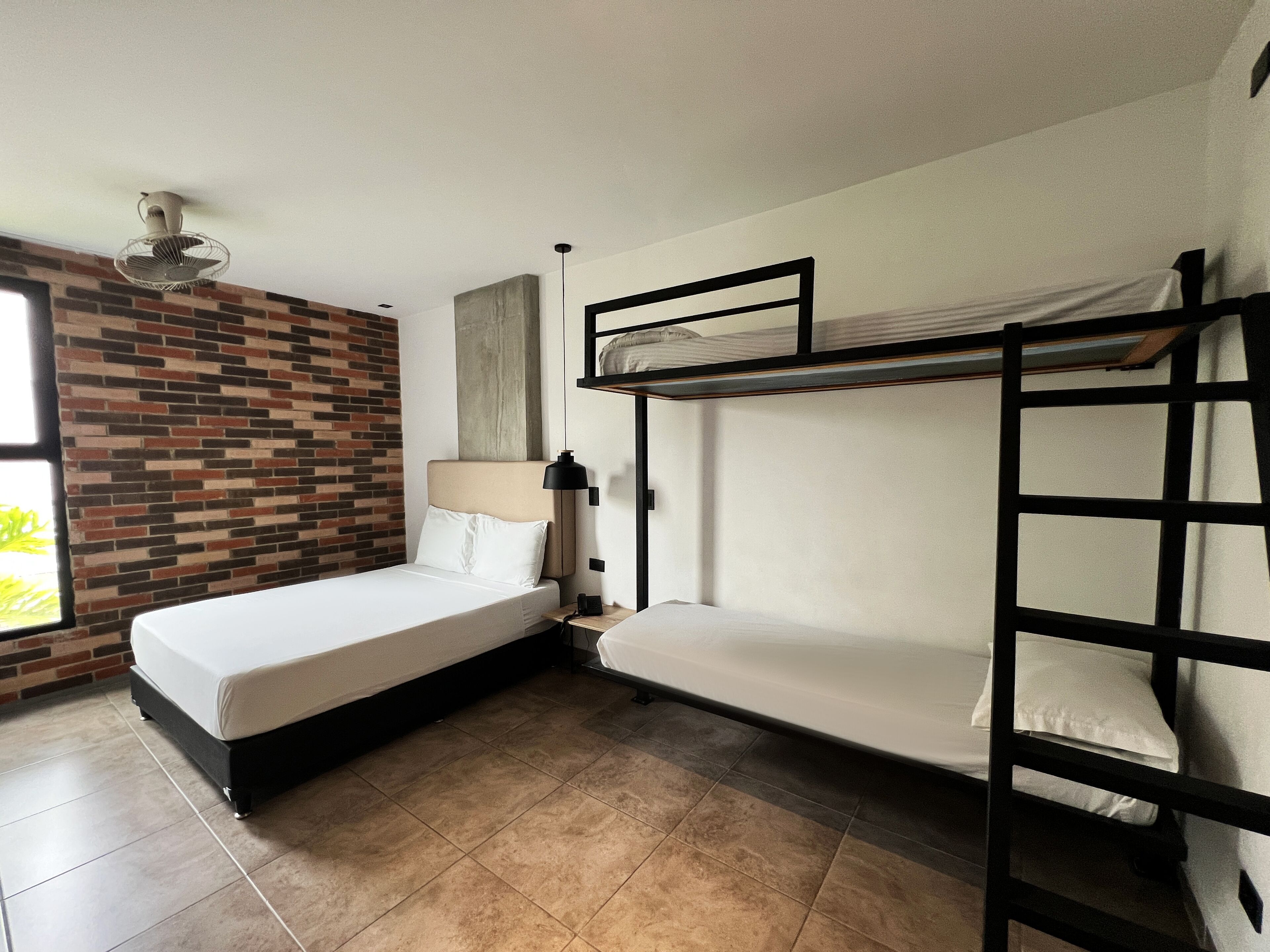Ein Zimmer mit einer Wand im Ziegelsteinmuster, einem Bett mit weißer Matratze und Kissen, einem Nachttisch mit Lampe und einem Stockbett mit weißer Matratze.