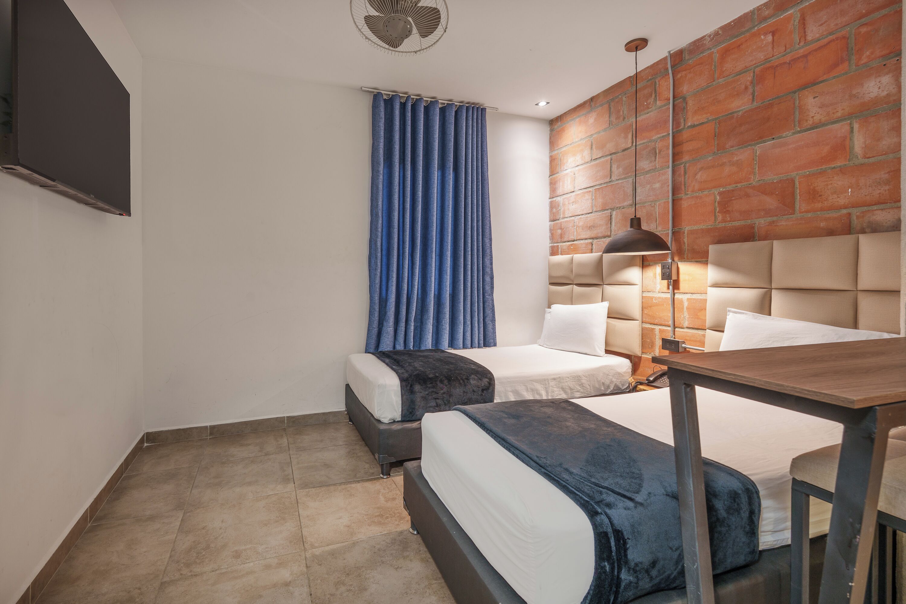 Una habitación de hotel con dos camas, una pared de ladrillo, un ventilador de techo y una mesa.