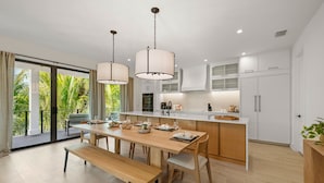 A modern kitchen with a wooden dining table, pendant lights, and a view of lush greenery outside.