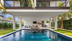 A modern outdoor pool area with a covered seating area, lounge chairs, and a flat-screen TV mounted on the wall.