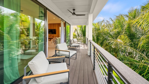 A balcony with white chairs and a ceiling fan, overlooking a lush garden with palm trees.