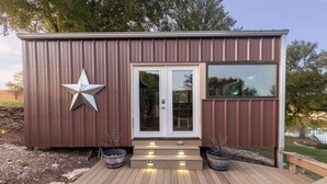 Un pequeño edificio metálico marrón con decoración de estrella, puerta de cristal y escalones que conducen a una terraza de madera.