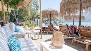 Un área de descanso frente al mar con sofás blancos, sillones de madera, sombrillas de paja y cestas tejidas sobre las mesas.