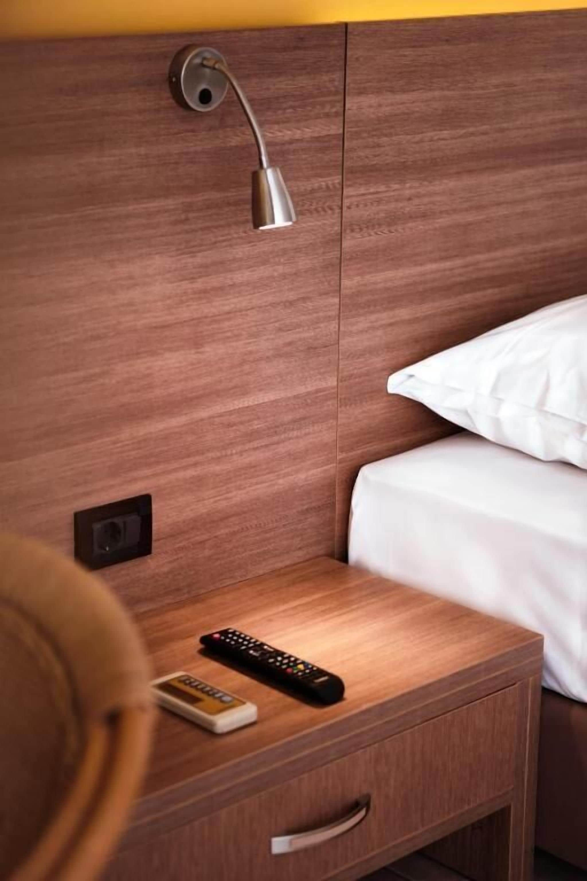 Una mesita de noche de madera con un control remoto y un pequeño dispositivo, una lámpara de pared y una cama con ropa blanca.