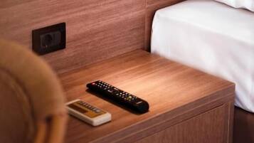 Una mesita de noche de madera con un control remoto y un pequeño dispositivo, una lámpara de pared y una cama con ropa blanca.
