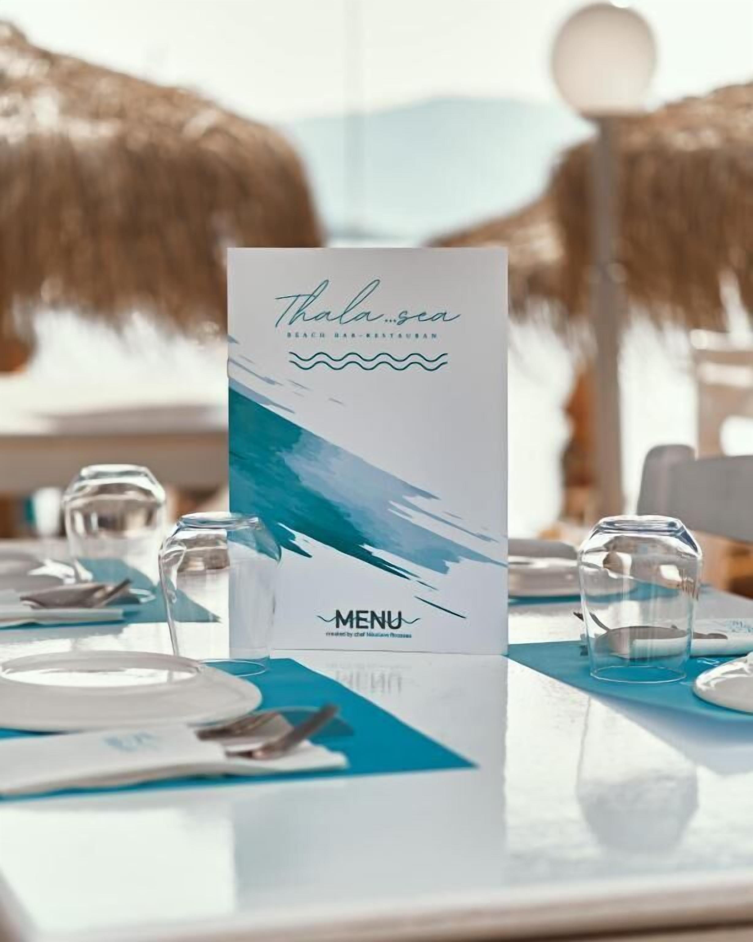 Un atril con menú, un letrero de restaurante de bar en la playa, una mesa con platos y vasos, y una sombrilla de paja al fondo.