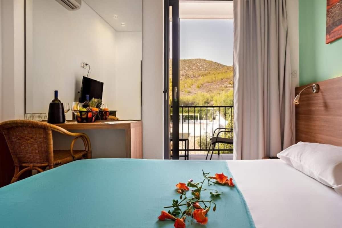 A hotel room with a bed, a desk with a computer, a chair, and a balcony with a view of a hilly landscape.