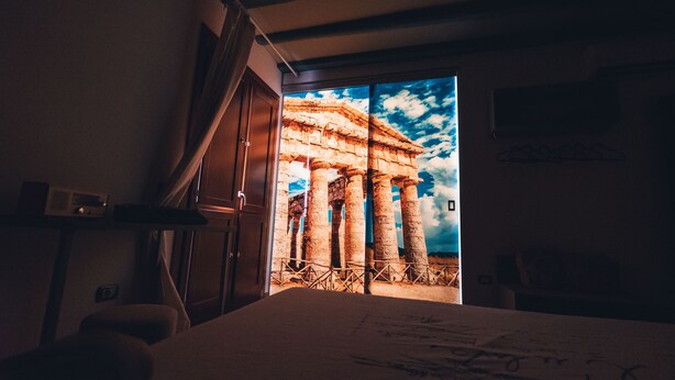 A bedroom with a large bed, a nightstand, and a window with a view of ancient columns. - macchiamediterrane (Castellammare del Golfo)