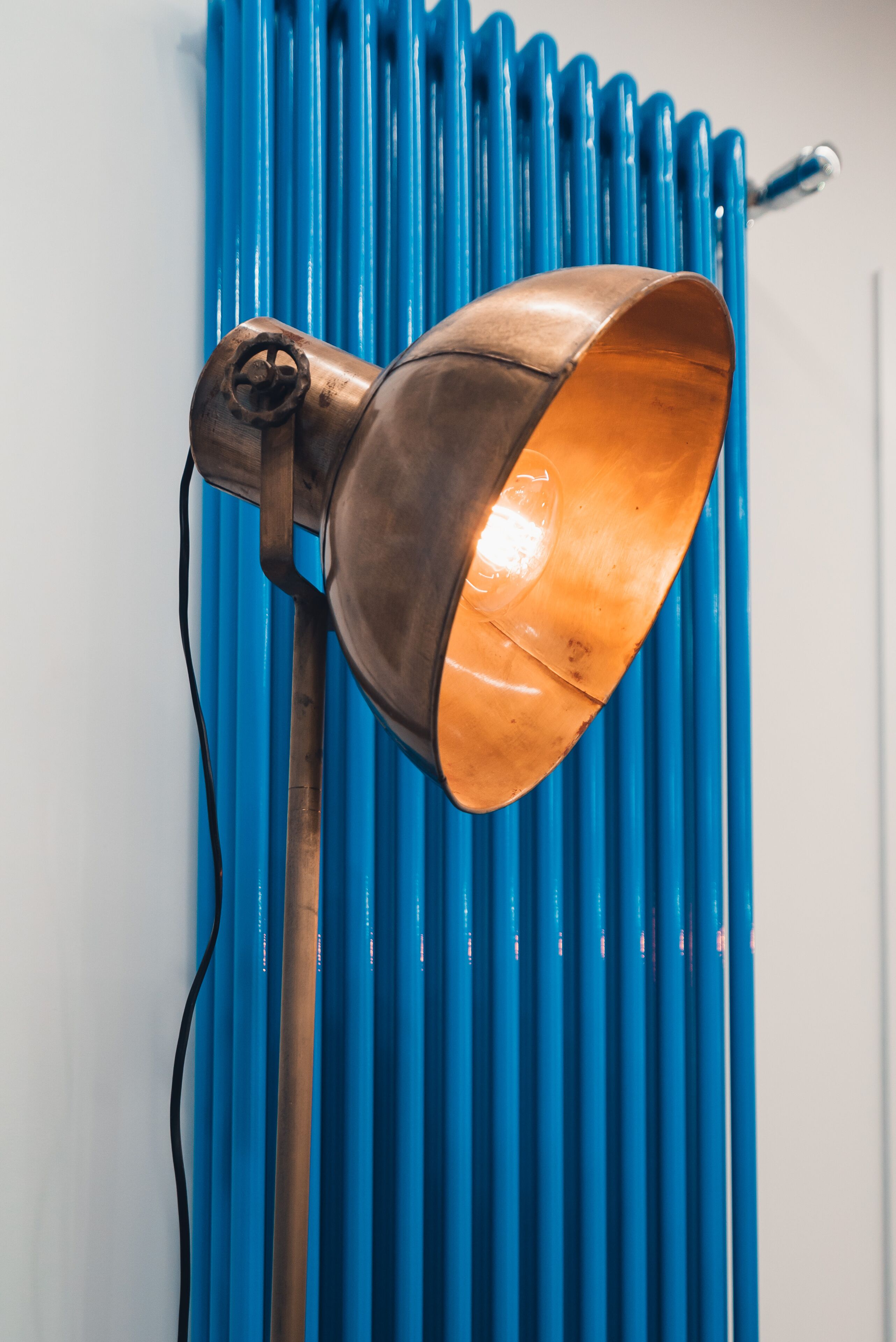 Una lámpara de pie color cobre con una pantalla curva frente a un radiador azul.