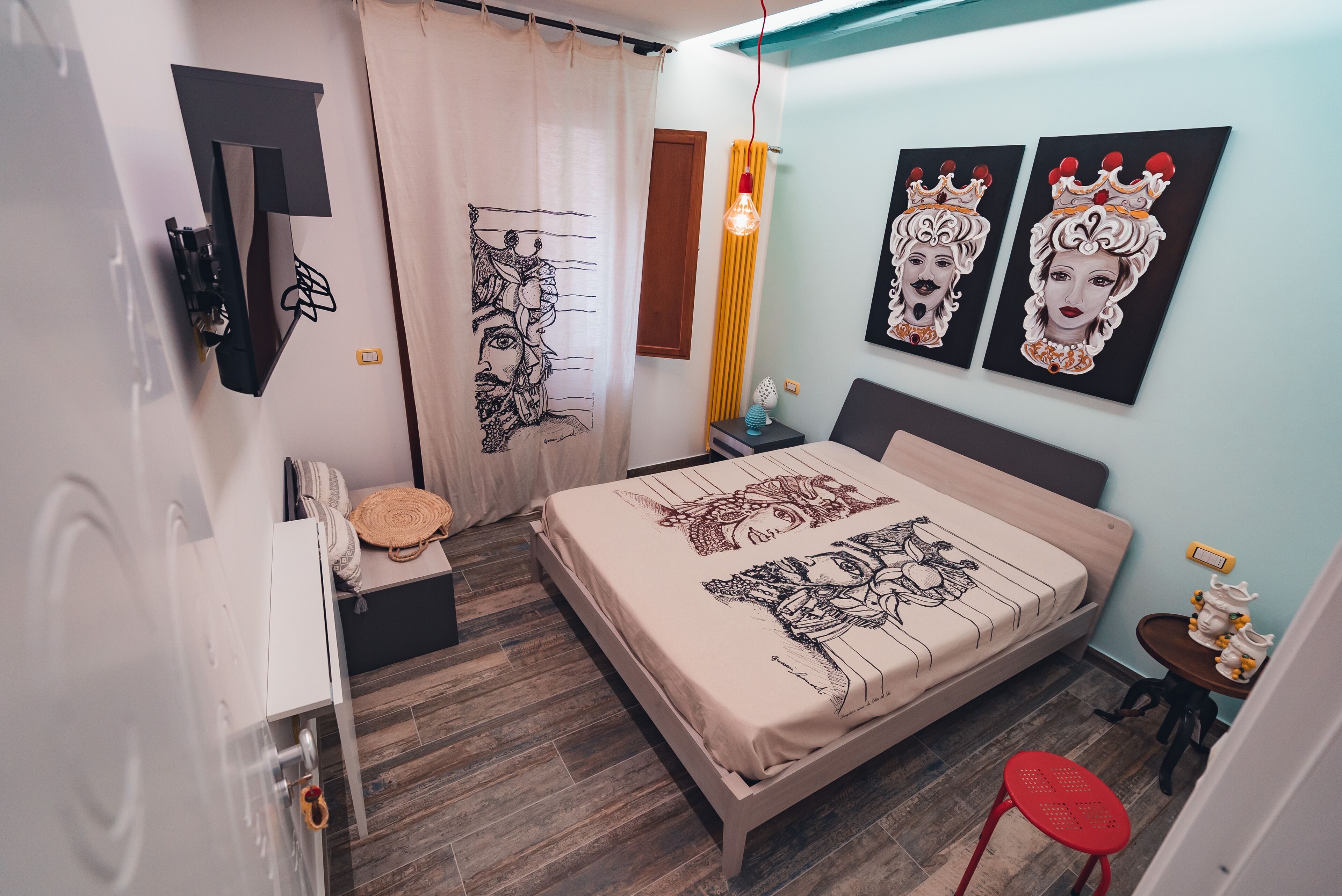 Un dormitorio con una cama, una mesita de noche, un taburete rojo, una lámpara de pared y dos cuadros.