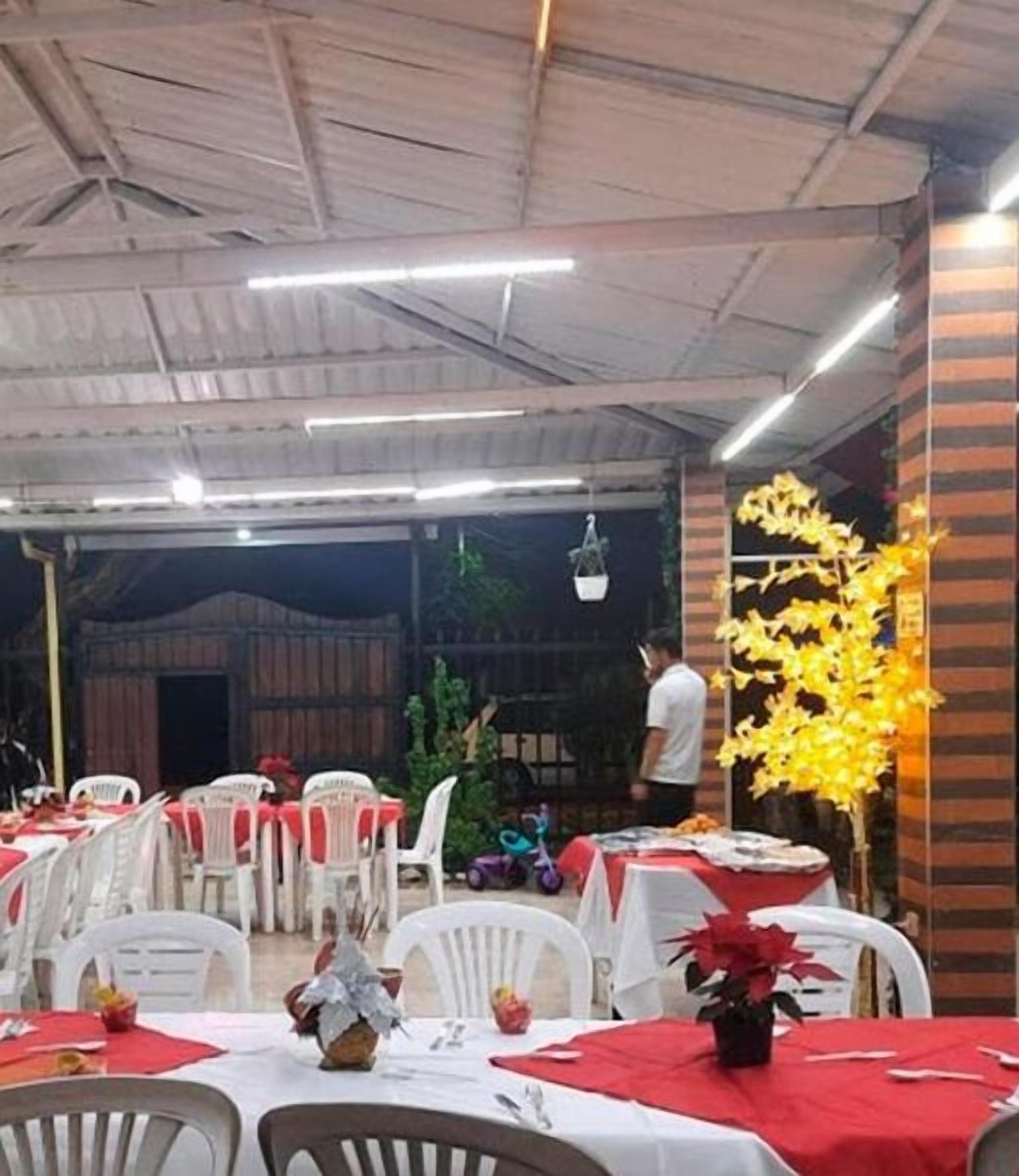 An indoor event space with tables covered in red tablecloths, white plastic chairs, and a person standing near a yellow decorative plant.