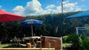 Un patio con una mesa de madera y bancos, rodeado por un seto y un paraguas rojo.