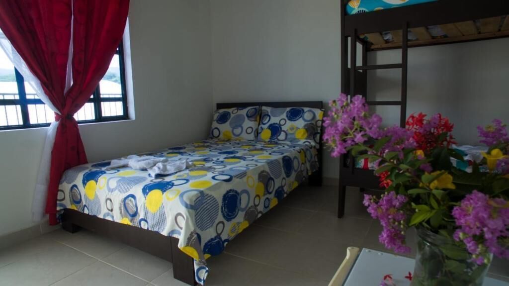 A single bed with a patterned bedspread, a bunk bed to the right, a window with red curtains, and a vase with flowers on a table.