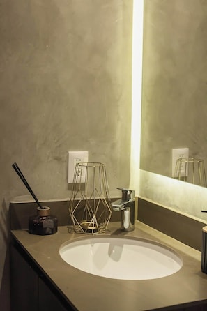 A bathroom with a round sink, a gold-colored geometric stool, a black bottle, and a silver pitcher.