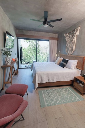 Une chambre moderne avec un grand lit, un ventilateur de plafond, une vue à travers une porte-fenêtre coulissante, une décoration murale en macramé et une table de chevet en bois.