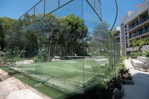 A tennis court enclosed by glass fencing, surrounded by greenery and modern buildings.