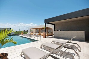 Un espace piscine sur le toit, doté de chaises longues, d’un espace salon fermé par des baies vitrées et d’une architecture moderne.