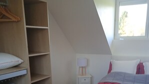 Ein Schlafzimmer mit schrägen Wänden, einem Bett mit roter Decke, einem Holzschrank, einem Nachttisch mit Lampe und einem Fenster mit Blick auf Bäume.