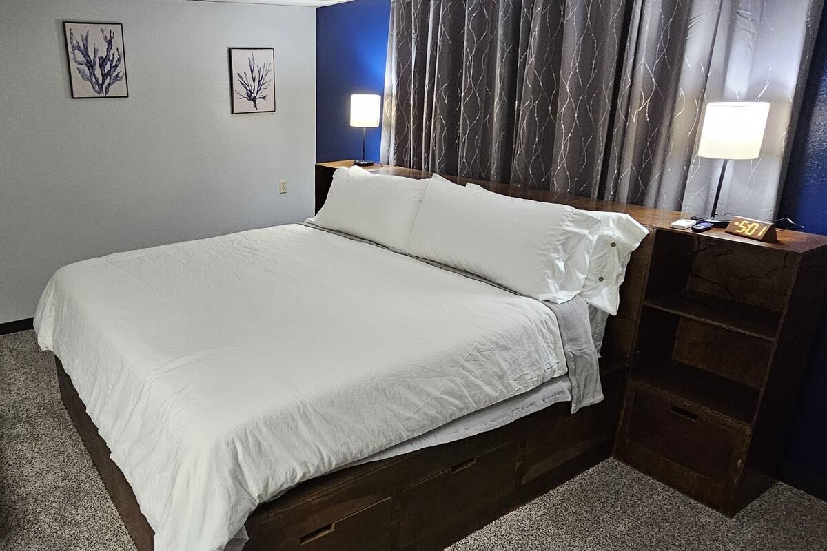 A neatly made bed with white linens, a wooden headboard, a nightstand with a lamp, and a small shelf.