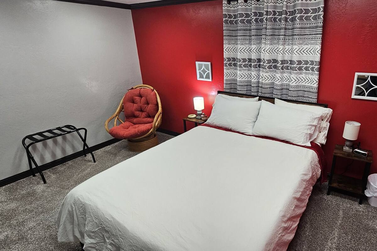 A bedroom with a bed, a chair, a bedside table with a lamp, and a wall with a patterned curtain.