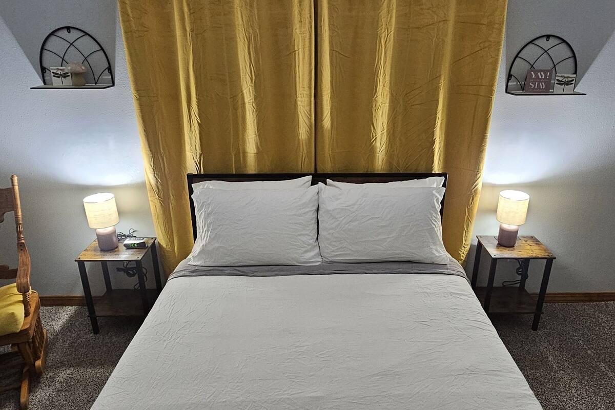 A neatly made bed with white linens, flanked by two bedside lamps, a wooden chair with a yellow cushion, and a small table with a clock.