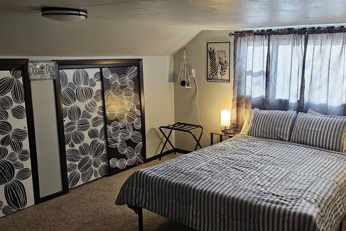 A bedroom with a bed, bedside lamps, a nightstand, a window with patterned curtains, and wall art.