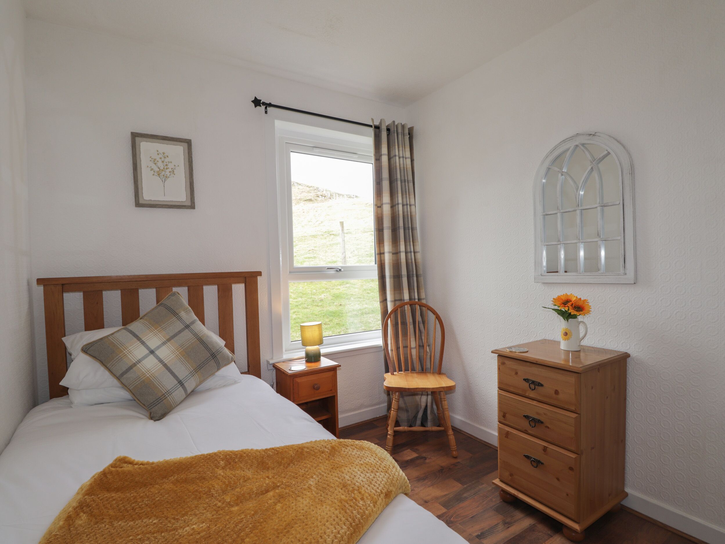 Une chambre à coucher comprenant un lit, une table de chevet en bois, une chaise, une commode, une fenêtre avec des rideaux et un miroir fixé au mur.